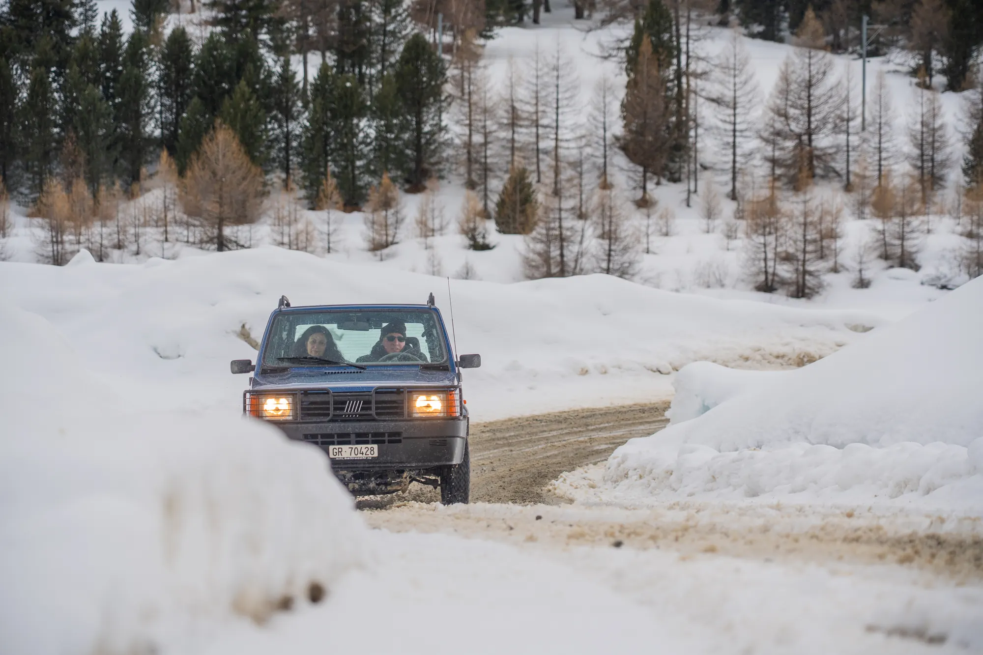 Fiat Panda 4x4 mit eingeschalteten Scheinwerfern auf einer verschneiten Piste vor Lärchen im Engadin