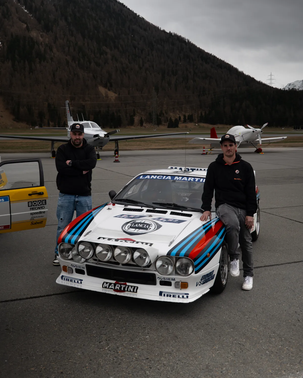 Fabio und Davide, die Gründer von Meet & Drive, mit einem Lancia Rally 037 in Martini-Farben auf einem Flugplatz im Engadin