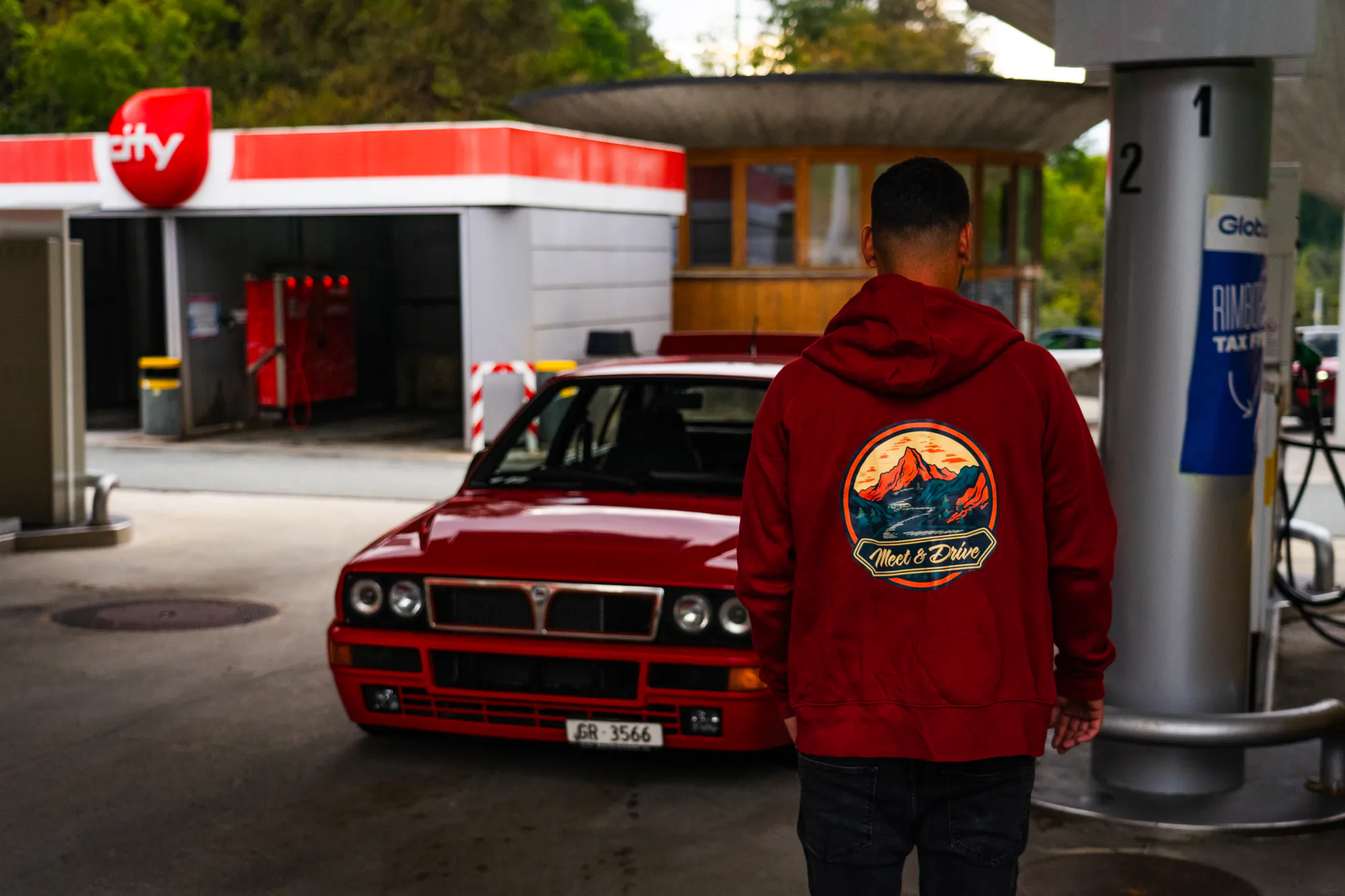 Community-Mitglied im Meet & Drive Hoodie neben einem roten Lancia Delta an der Tankstelle
